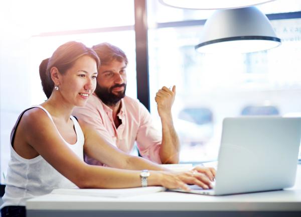 A man and a woman sit in a bright office space, working on a laptop to discover how to teach online university courses